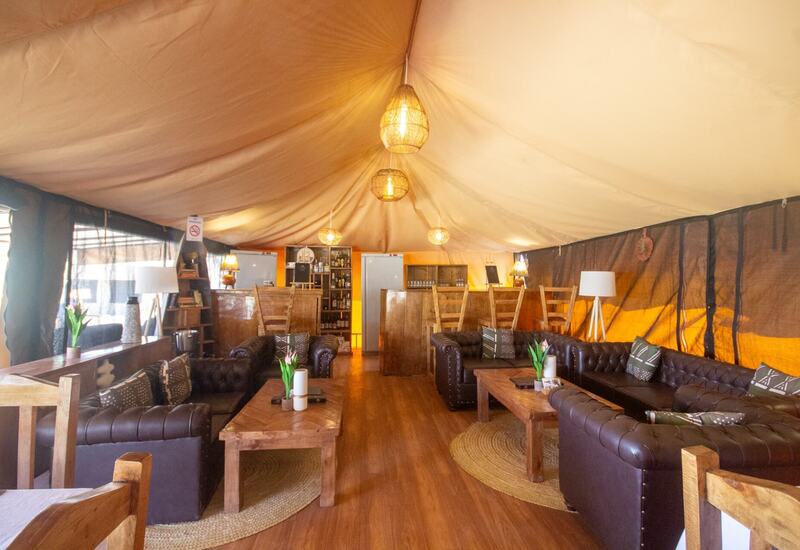 Spacious safari-style lounge tent with leather chesterfield sofas, wooden coffee tables, woven pendant lights and a stocked bar area