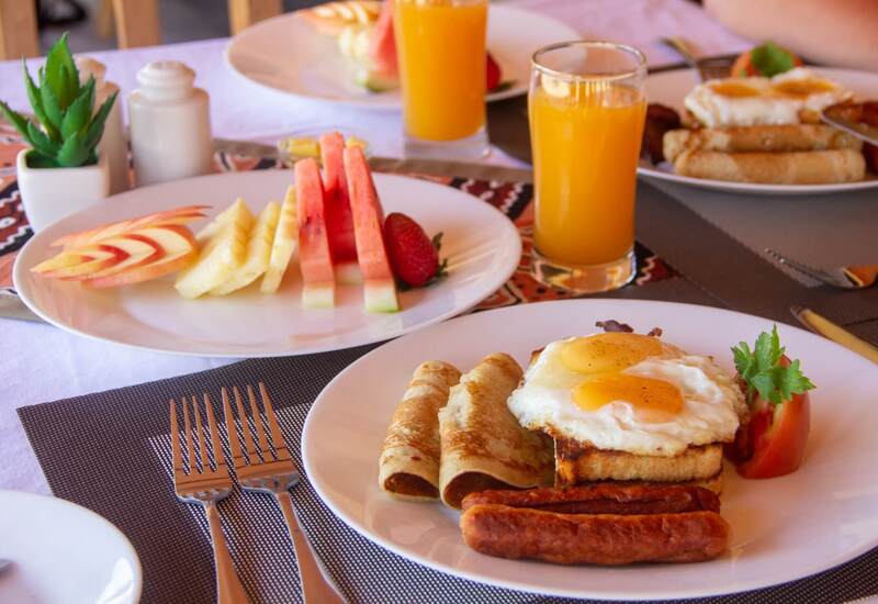 Hearty hotel breakfast with fried eggs on toast, sausages and rolled crepes, fresh fruit slices and glasses of orange juice