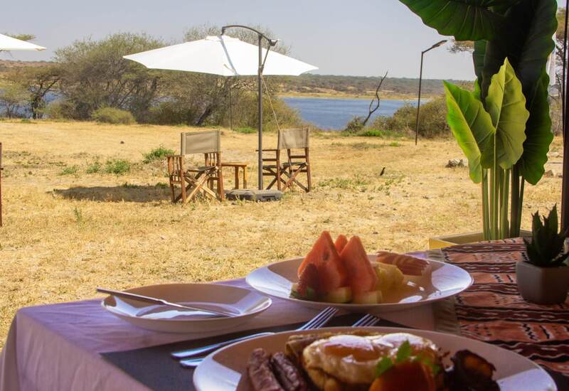 Al-fresco breakfast plate on a table framed by tropical leaves, facing wooden chairs under a white umbrella by a river.