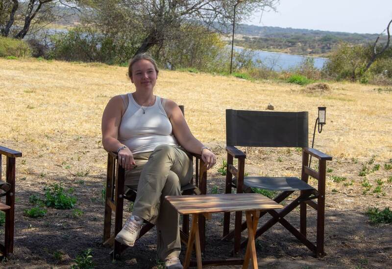 Woman relaxing in a canvas chair at a shaded lakeside campsite, wooden table, dry grass and trees by a calm water view