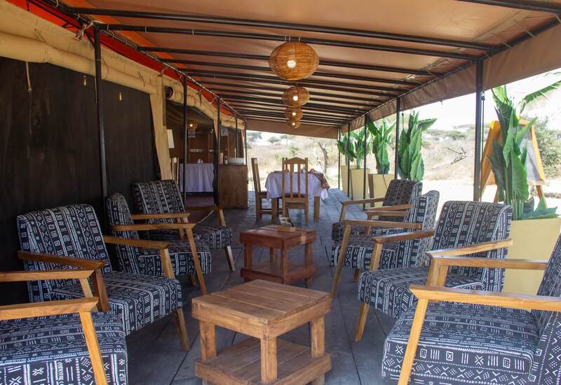 Safari tent lounge with wooden armchairs in patterned fabric, woven pendant lamps, dining table and open savanna view.