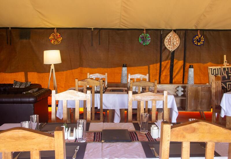 Tented camp dining with rustic wooden chairs, linen-covered tables, warm amber lighting, leather sofa and wall decorations.