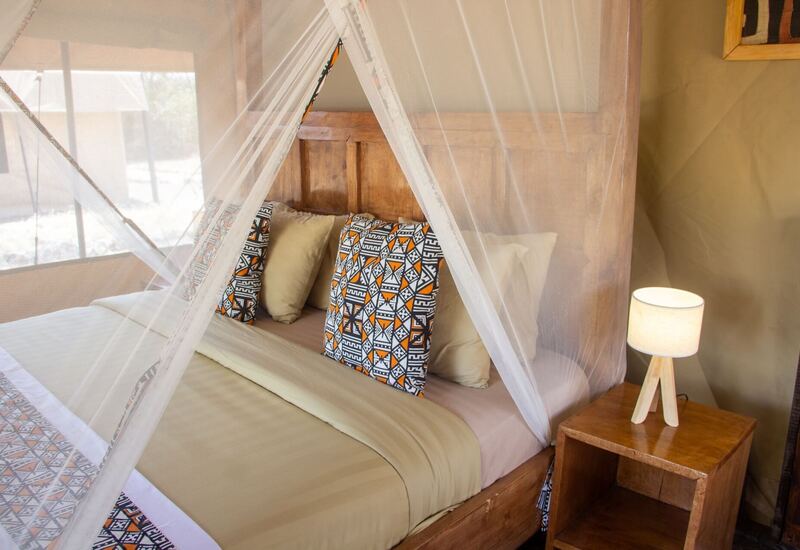 Safari-style tent bedroom with wooden headboard, sheer mosquito-net canopy, geometric orange-black pillows and wooden bedside lamp.