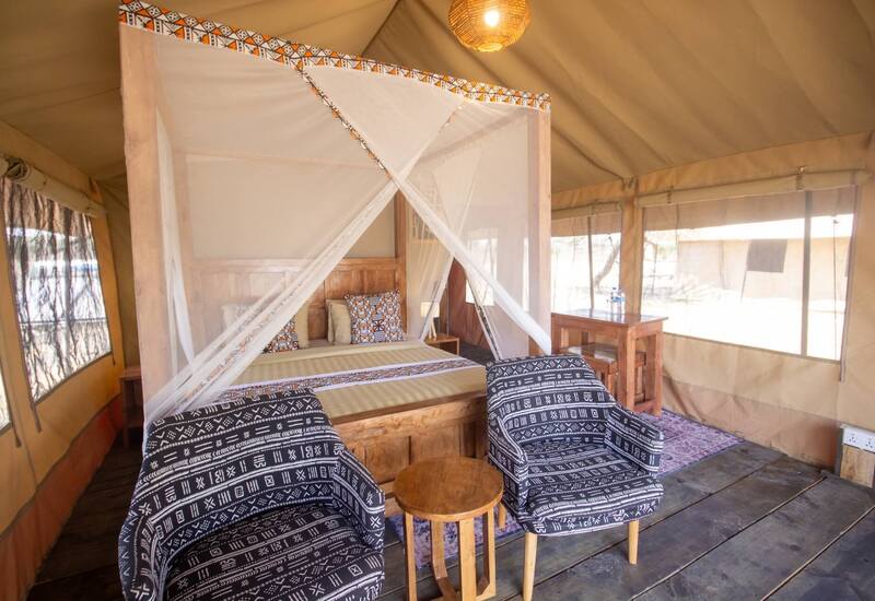 Tented safari suite with four‑poster bed draped in mosquito net, patterned pillows, wooden furnishings and two patterned armchairs.