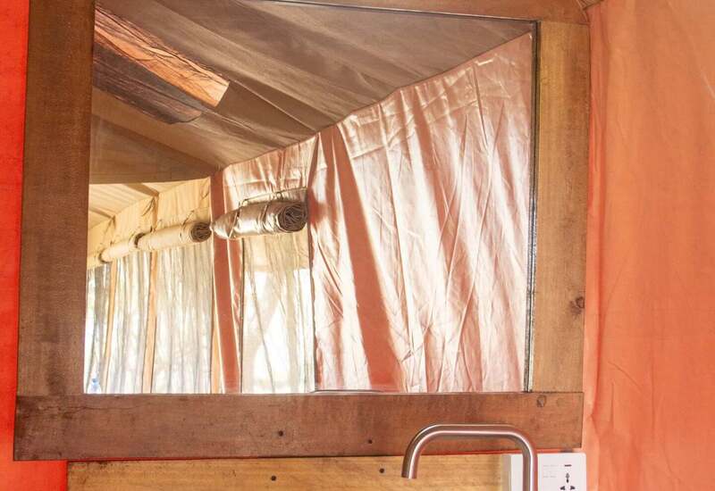 Wood-framed mirror and light above a white vessel sink with stainless faucet, soap and folded towels in orange canvas tent.