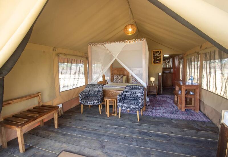 Spacious safari tent with four-poster bed and mosquito net, patterned cushions, two armchairs, bedside lamp and small desk.