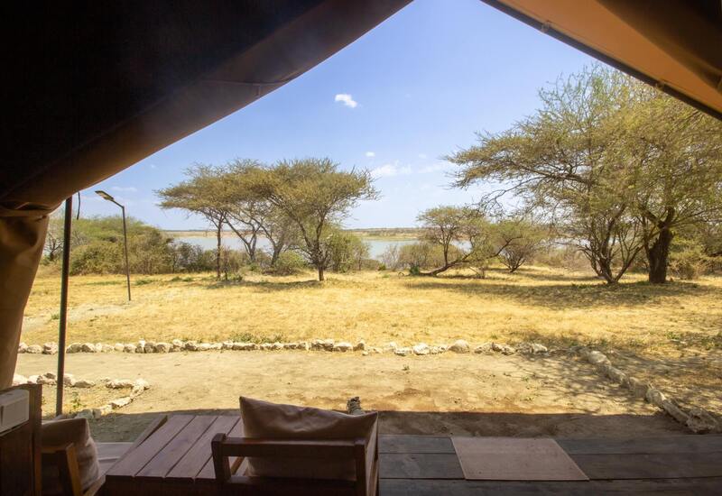 Tented safari lodge veranda with wooden deck and cushions overlooking dry grass, acacia trees and a tranquil lake view.