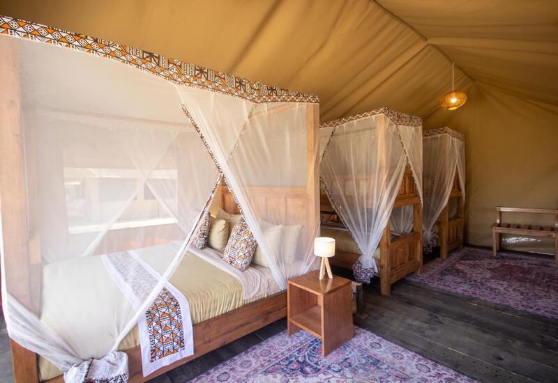 Tented glamping room with wooden four-poster beds draped in white mosquito nets, patterned trim, bedside lamp and rugs.