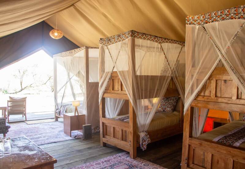 Luxury safari tent with wooden bunk beds draped in mosquito nets, patterned textiles, warm lantern light and open deck view.
