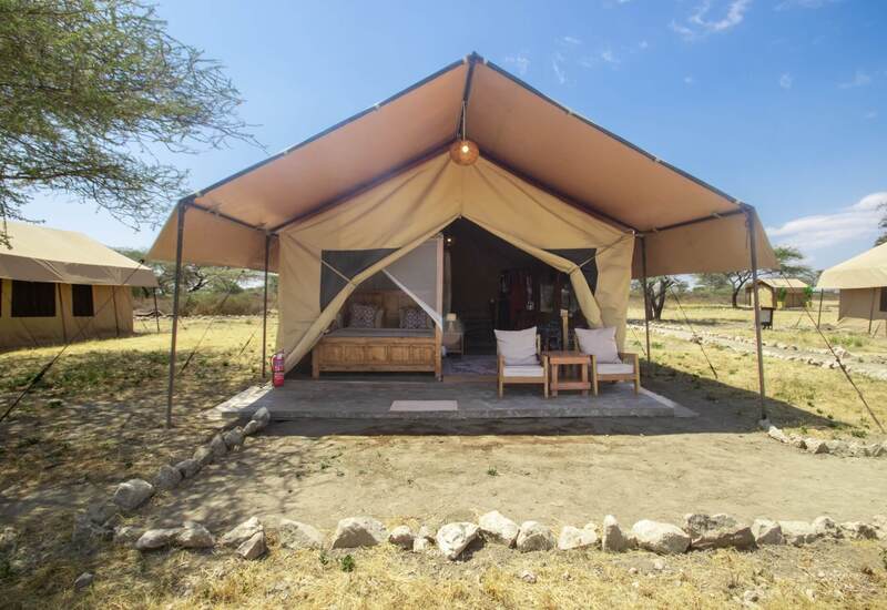 Canvas safari tent with shaded wooden porch, king bed with mosquito net, two cushioned chairs, lantern and dry savannah view.