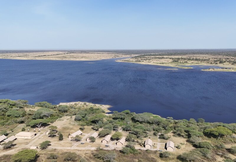 Lakeside safari camp with canvas tents along a deep blue river, sandy banks, acacia trees and wide open savanna.