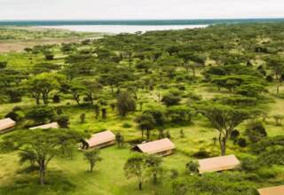 Tented safari cabins scattered among acacia trees on a green savanna, overlooking a distant lake, secluded safari camp setting.
