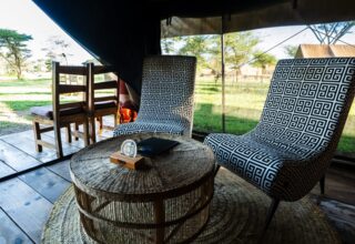 Tented safari lounge with two patterned chairs, woven round table and wooden deck overlooking savanna and a parked jeep.