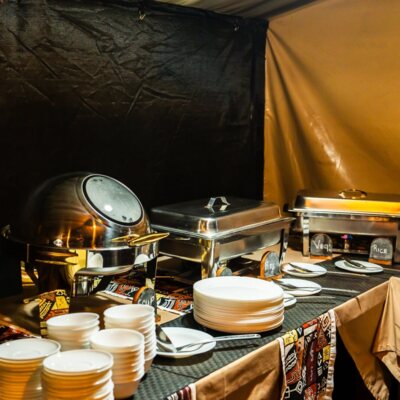 Warm tented buffet with stainless chafing dishes, labeled trays (rice, veg), stacked plates and bowls and serving spoons on patterned tablecloth.