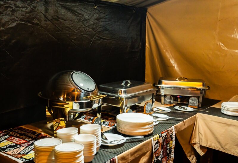 Warm tented buffet with stainless chafing dishes, labeled trays (rice, veg), stacked plates and bowls and serving spoons on patterned tablecloth.