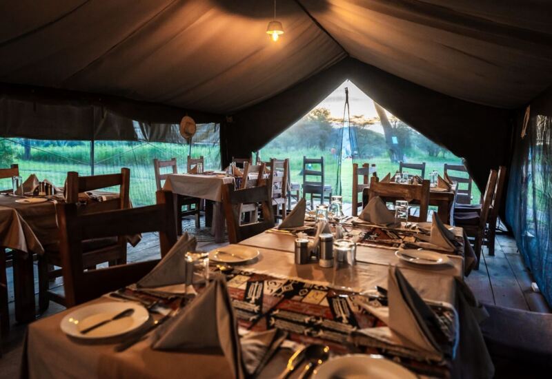 Safari dining tent with wooden tables set for dinner, folded napkins and cutlery, warm light and grassy savanna views.