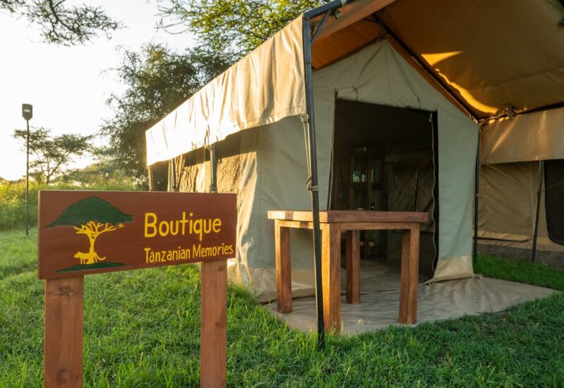Boutique Tanzanian Memories sign by canvas safari tent shop with wooden table on grassy campsite at golden hour
