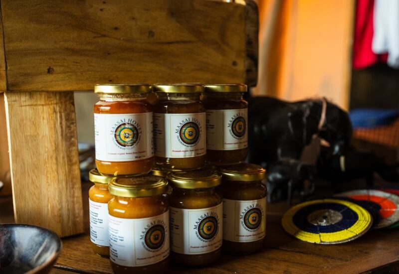 Stack of artisanal honey jars labeled 'Maasai Honey' on wooden shelf beside carved elephant and colorful beaded coasters.