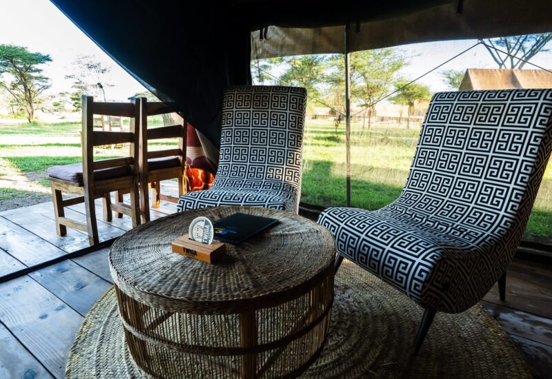 Tented safari lounge with two patterned chairs, woven round table and wooden deck overlooking savanna and a parked jeep.