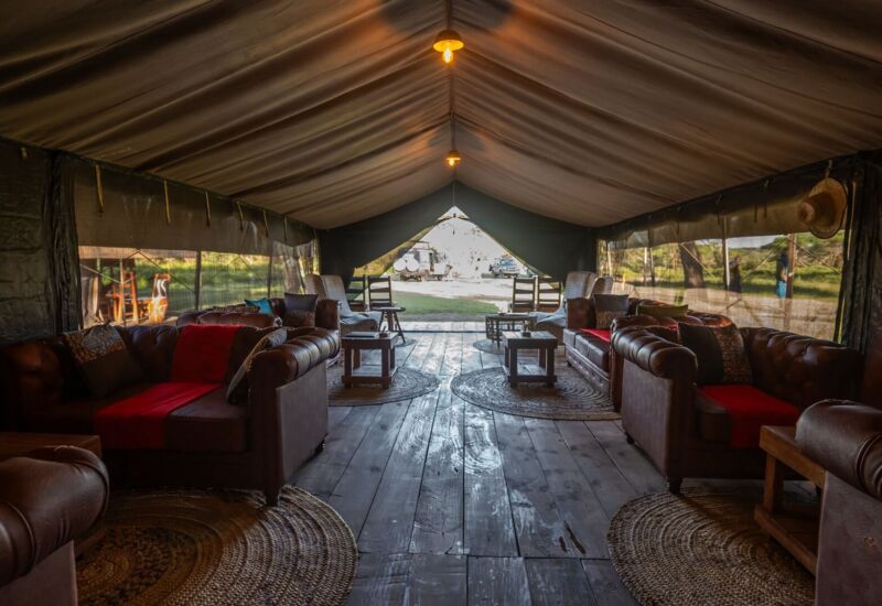 Spacious luxury safari tent lounge with leather sofas, woven rugs, wooden floor, warm lighting and open savanna view