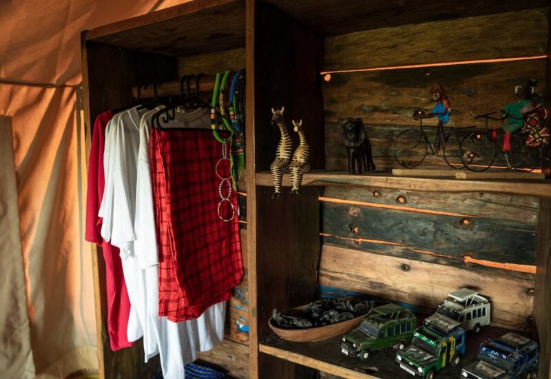 Canvas tent wardrobe with hanging shirts and red shuka, wooden shelves with beaded necklaces, carved zebras and toy jeeps