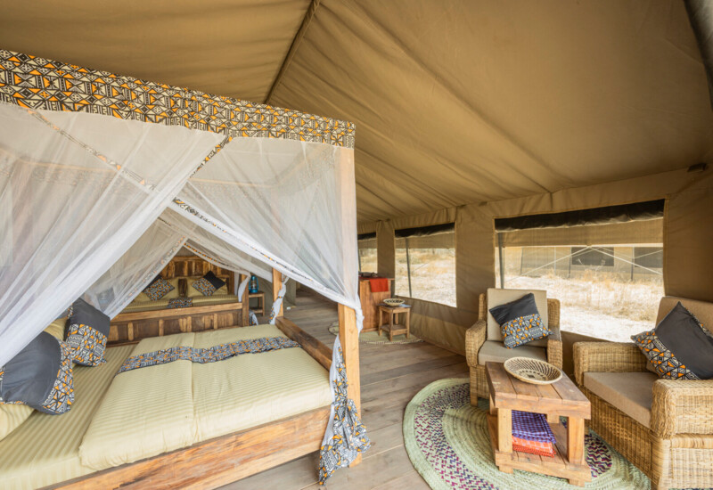 Spacious luxury safari tent with four-poster bed draped in mosquito net, wooden furnishings, rattan seating and savanna views.