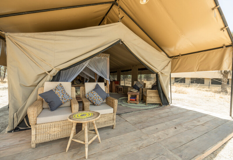 Safari glamping tent on wooden deck with wicker lounge chairs, patterned cushions and a mosquito-net bed visible inside.