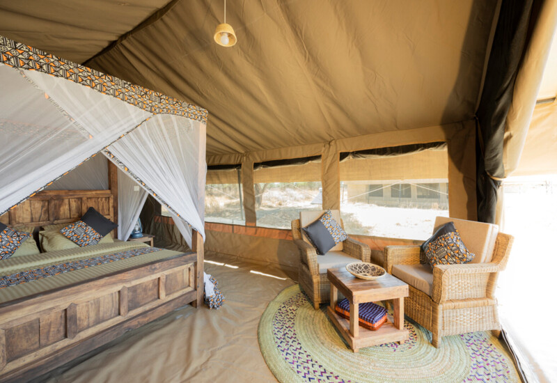 Spacious safari tent with wooden four-poster bed under mosquito net, wicker armchairs, coffee table and canvas windows.