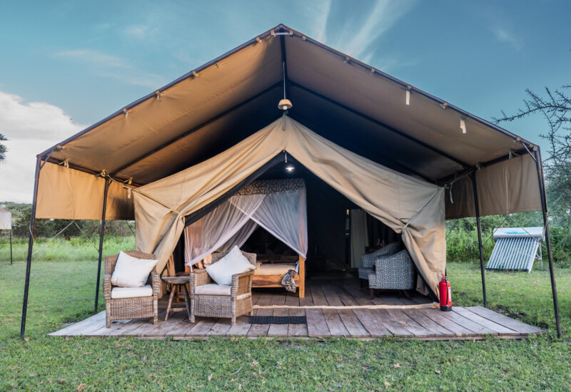 Luxury safari tent with wooden deck, wicker chairs and cushions, mosquito-netted bed inside, grassy savanna views.