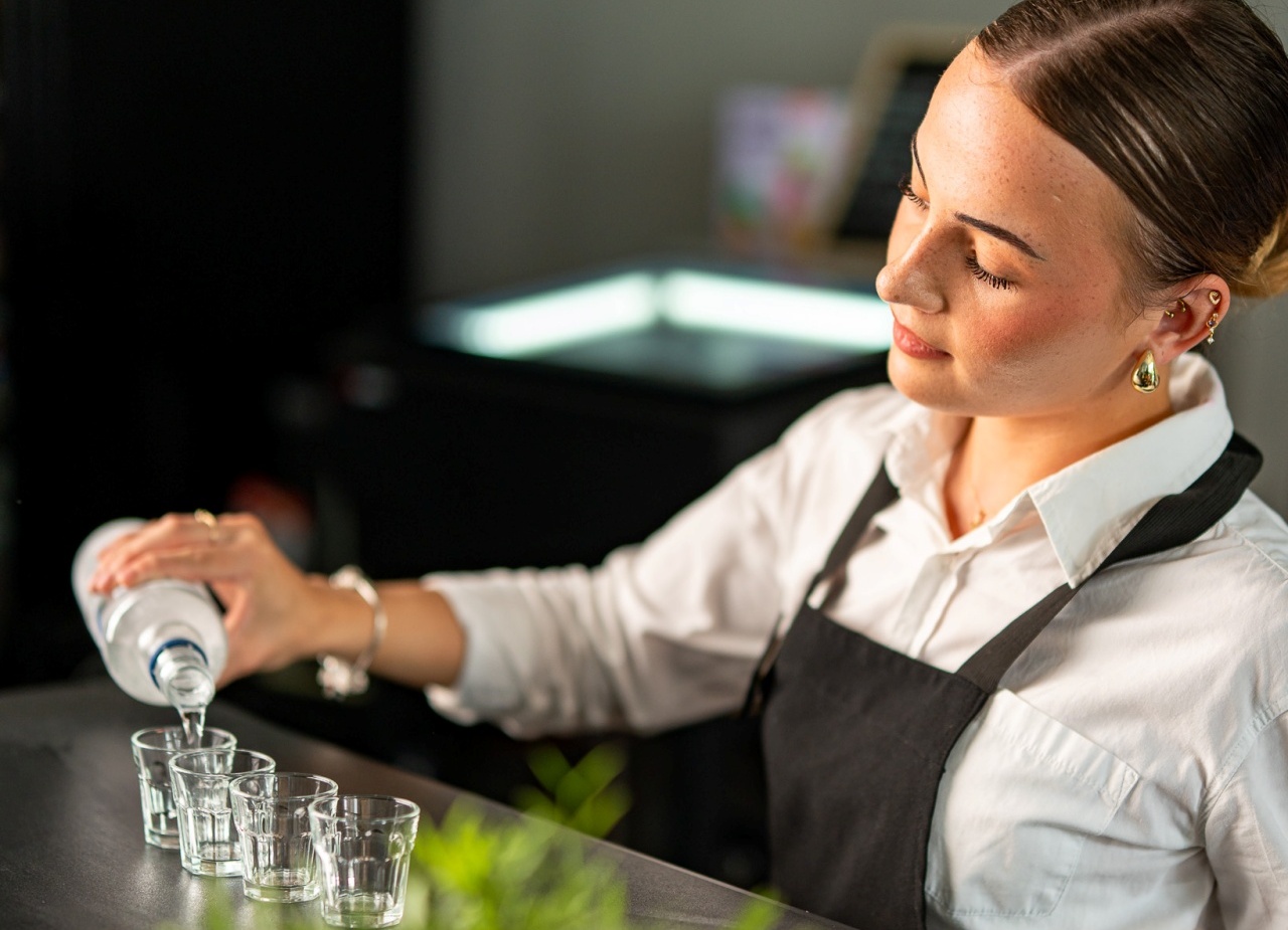 Barmanka w białej koszuli i czarnym fartuchu nalewa alkohol do czterech kieliszków na nowoczesnym barze