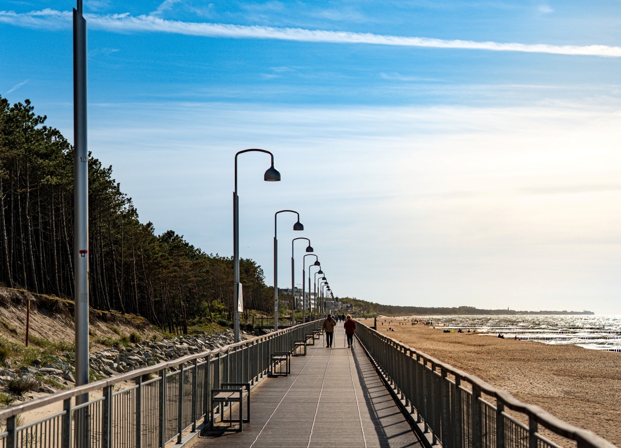Promenada nadmorska z rzędami latarń i ławek, piaszczysta plaża i morze, para spacerująca w oddali.