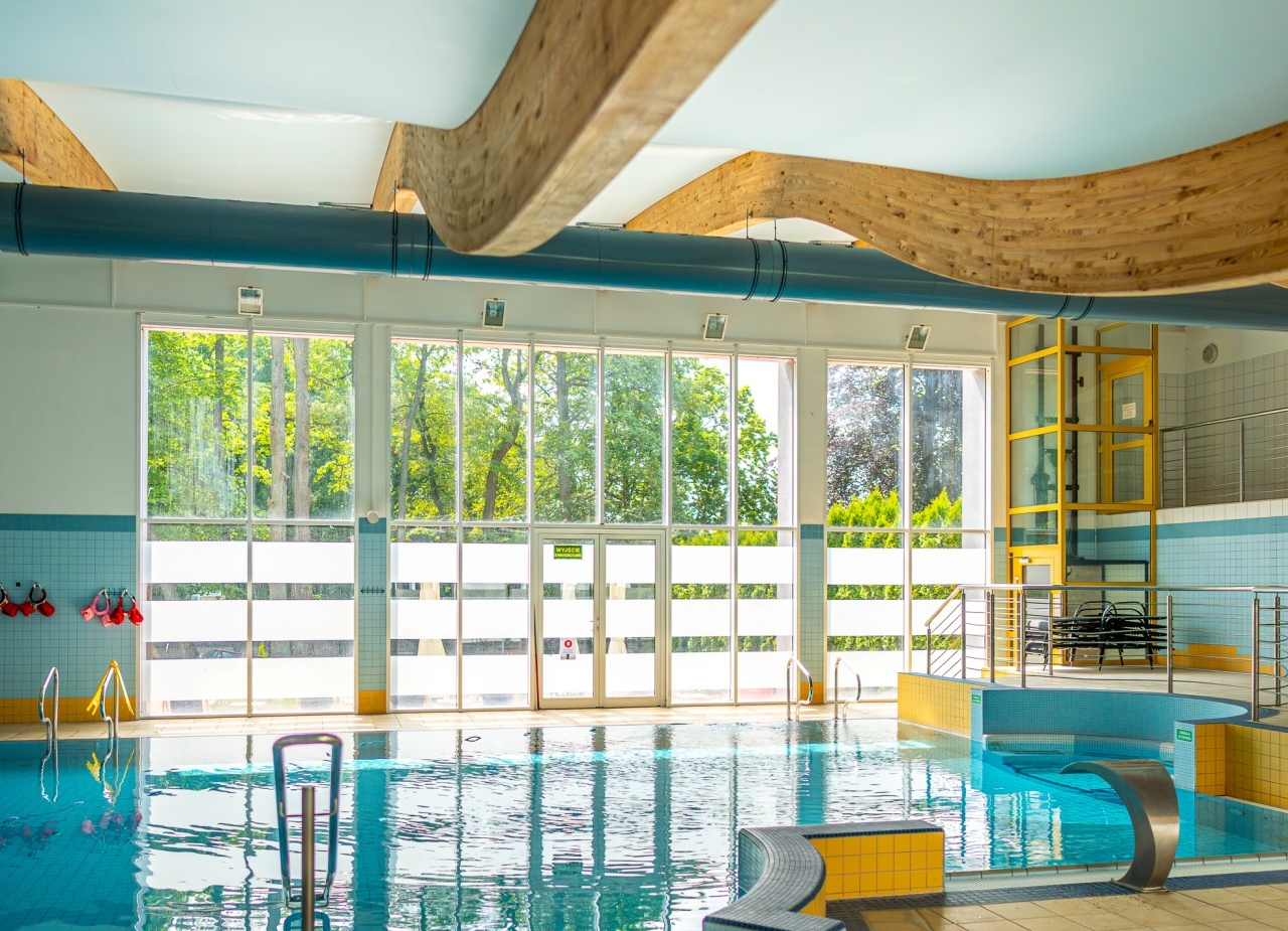 Wewnętrzny basen hotelowy z panoramicznymi oknami na zieleń, falistymi drewnianymi belkami, schodkami, metalową dyszą wodną i kaflowym tarasem