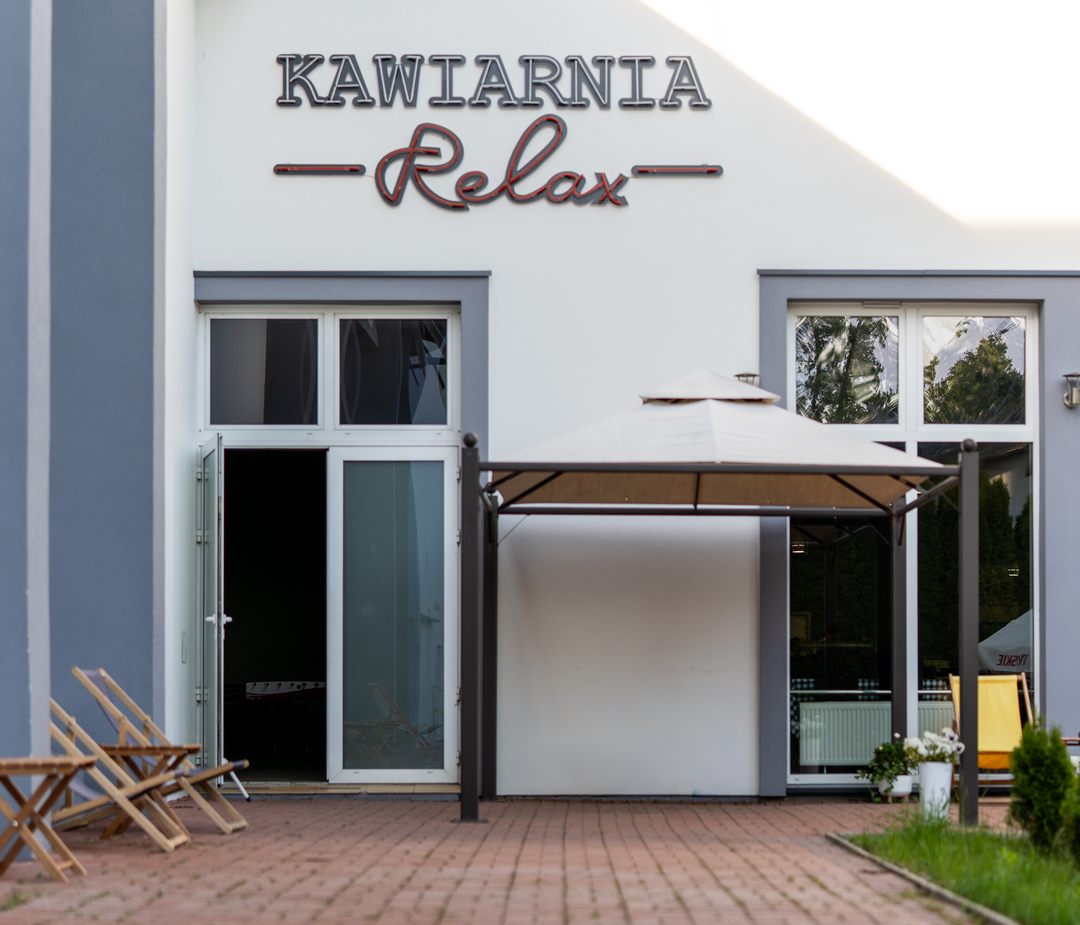 Kawiarnia Relax — biała elewacja z napisem, przeszklone drzwi, zadaszony taras, drewniane leżaki na ceglanej nawierzchni