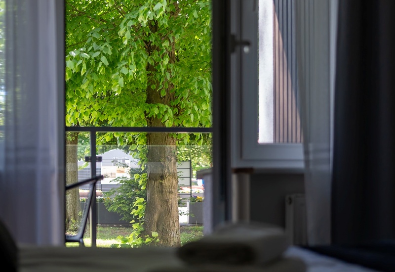 Pokój hotelowy z otwartym oknem na balkon, zasłony, ręczniki na łóżku i bujna zieleń drzew za szybą