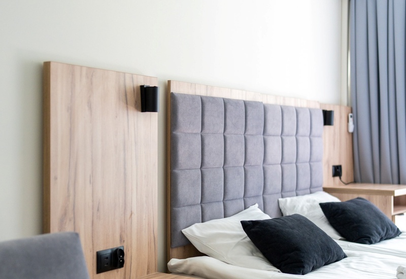 Nowoczesny pokój hotelowy z podwójnym łóżkiem, pikowanym szarym zagłówkiem, drewnianymi stolikami nocnymi, poduszkami, klimatyzacją i zasłonami.