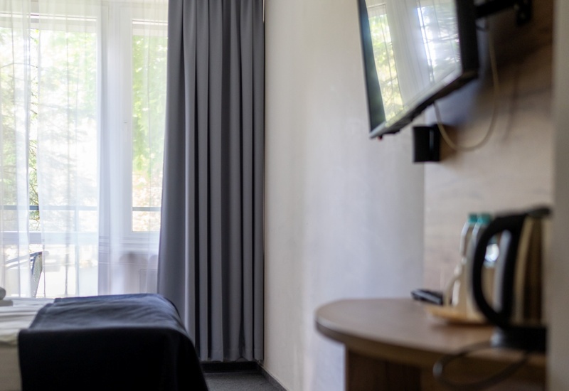 Pokój hotelowy z łóżkiem przy dużym oknie i balkonem za zasłonami, ścienny telewizor i czajnik na stoliku.
