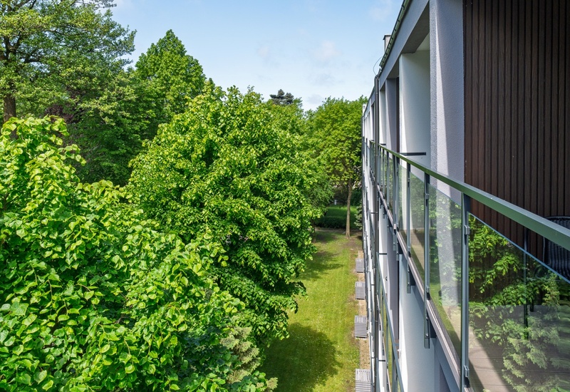 Nowoczesny balkon hotelu ze szklanymi balustradami, widok na gęsty zieleniec i prywatne tarasy przy trawiastym podwórzu