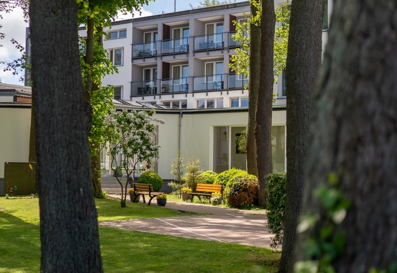 Cichy hotelowy dziedziniec z balkonami pokoi, zielonym ogrodem, ławkami i drzewami idealny do relaksu.