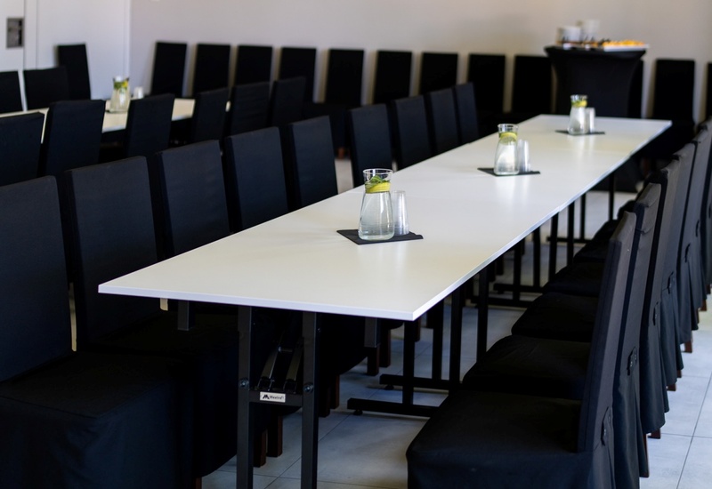 Sala konferencyjna z długimi białymi stołami, rzędem czarnych krzeseł, karafkami wody z cytryną i bufetem w tle.