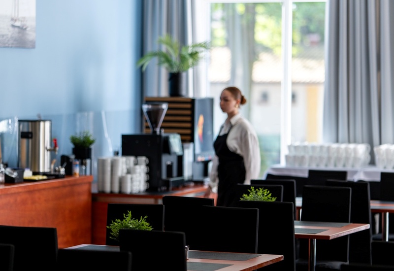 Restauracja hotelowa z kawowym barem, ekspresami, stolikami i roślinami; kelnerka przy dużym oknie.