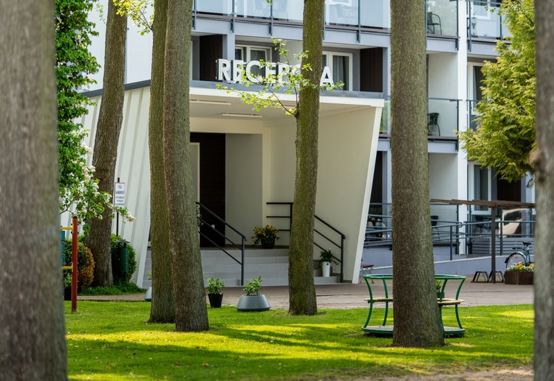 Wejście do recepcji nowoczesnego hotelu z przeszklonymi balkonami, schodami i donicami, w zielonym, parkowym otoczeniu