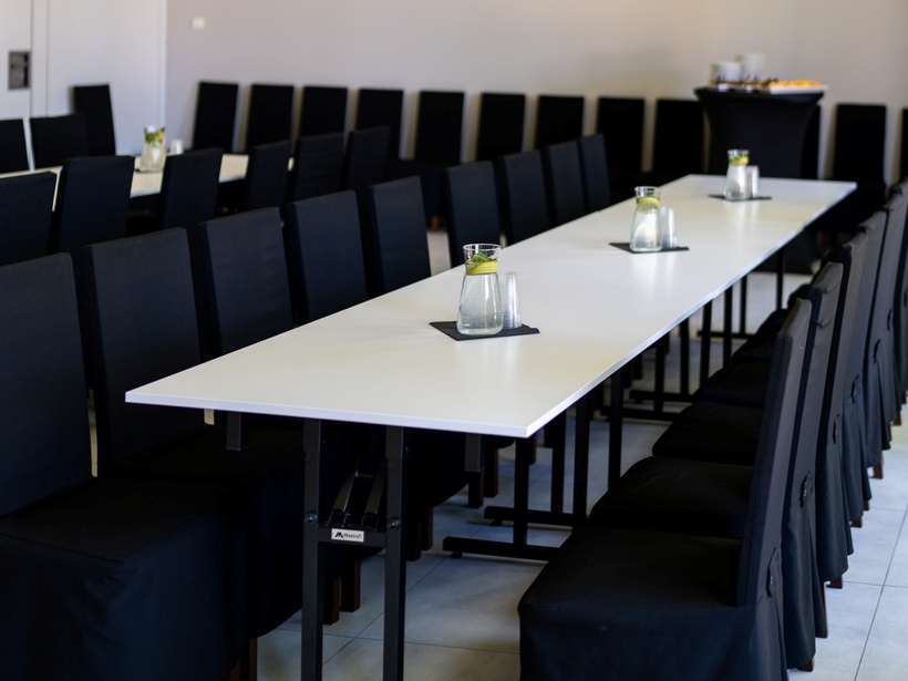 Sala konferencyjna z długimi białymi stołami, rzędem czarnych krzeseł, karafkami wody z cytryną i bufetem w tle.