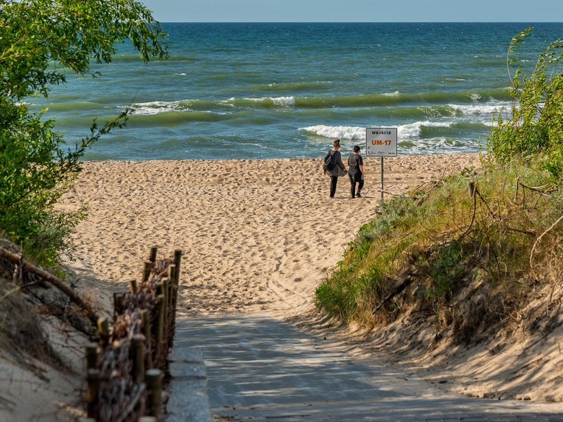 Ścieżka przez wydmy do piaszczystej plaży i falującego morza, przy znaku wejścia UM-17 dwie osoby idą ku wodzie.