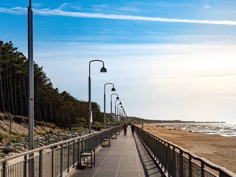 Promenada nadmorska z rzędami latarń i ławek, piaszczysta plaża i morze, para spacerująca w oddali.