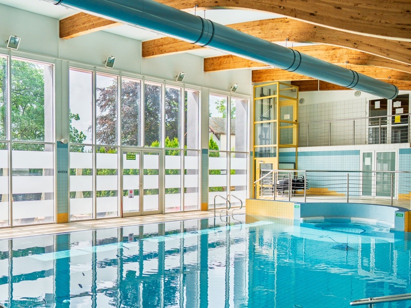 Kryty basen hotelowy z dużymi oknami, falistymi drewnianymi belkami, niebieskim kanałem i wejściem do wody.