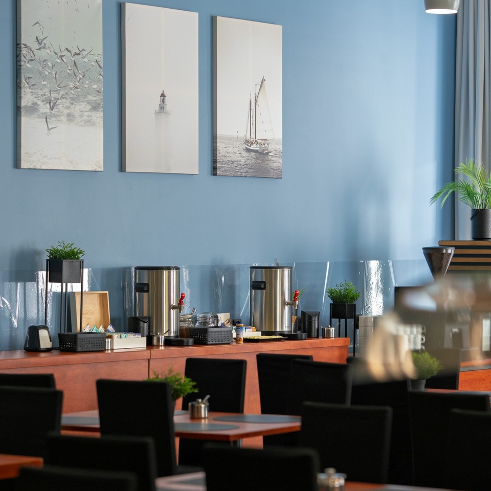 Jadalnia hotelowa z bufetem śniadaniowym, stalowymi podgrzewaczami kawy, drewnianymi stołami, czarnymi krzesłami i morskimi obrazami na niebieskiej ścianie