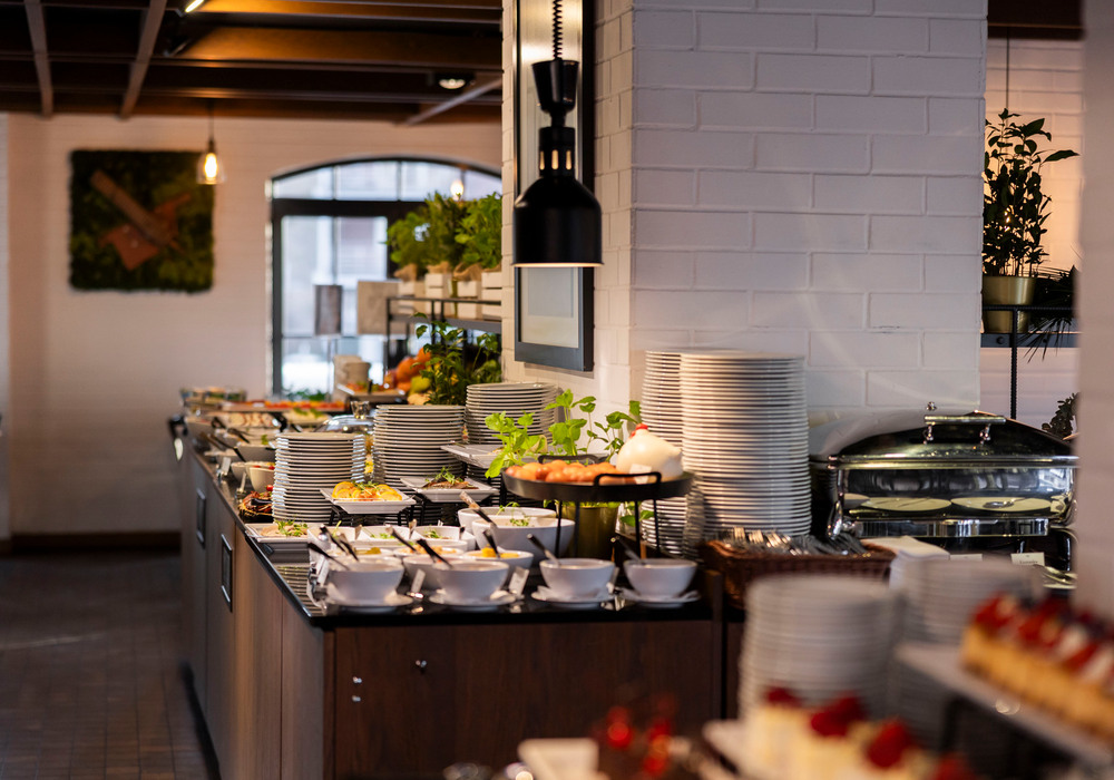 Buffet spread with stacked plates, bowls, chafing dish and salads on a wooden counter beneath warm pendant lights.