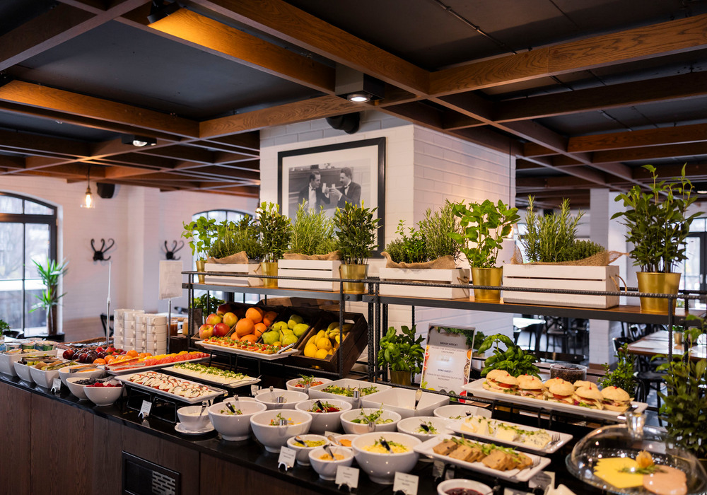 Hotel breakfast buffet with bowls of salads and fruit, slider sandwiches, fresh herbs on shelves, warm wood beams