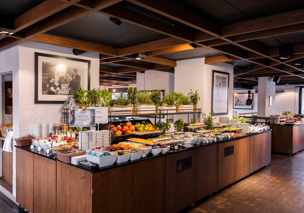 Spacious hotel breakfast buffet with fresh fruit, pastries, cereals, yogurt, coffee station, potted herbs and warm wood-beam ceiling.