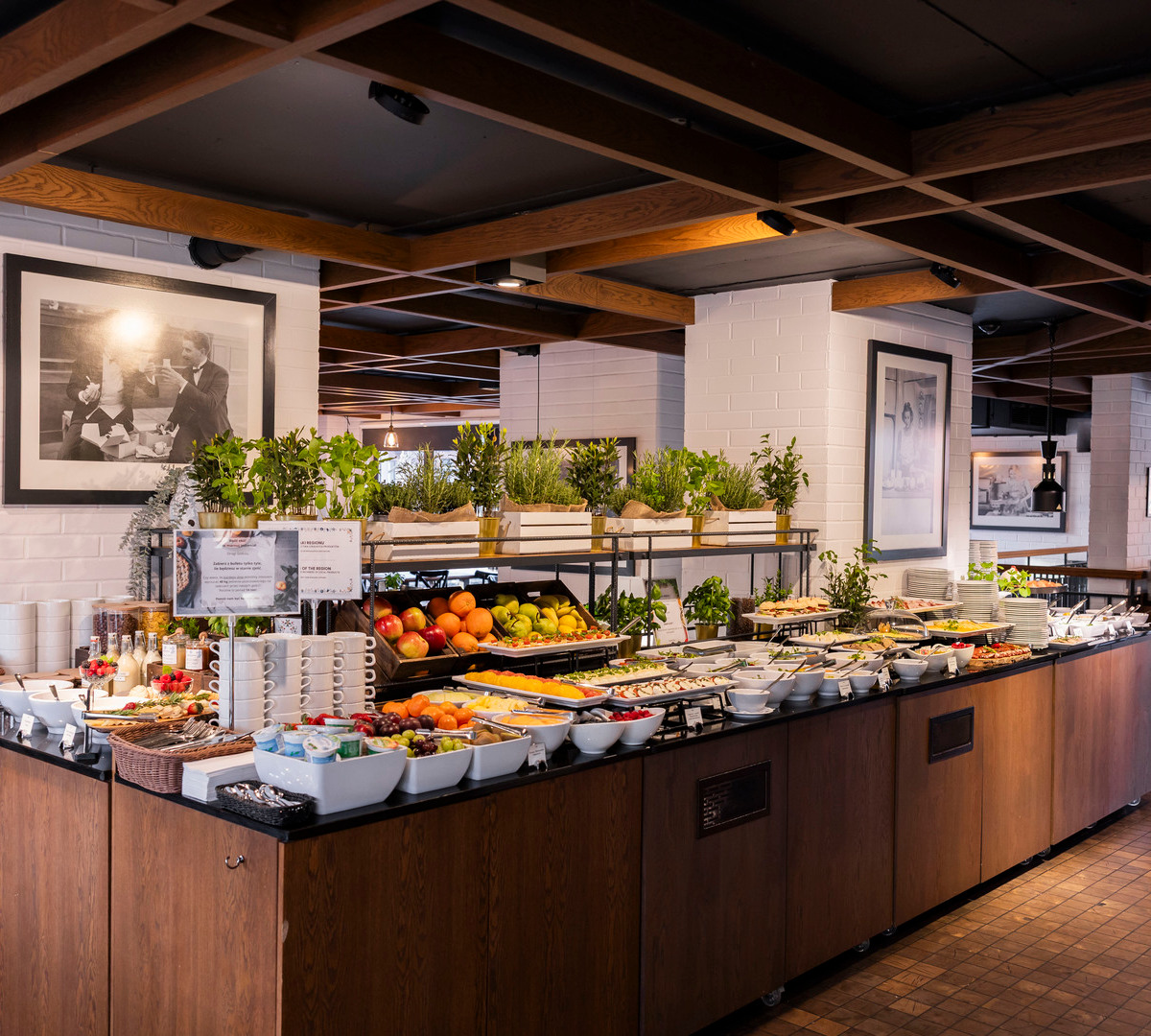 Hotellfrukostbuffé med färsk frukt, yoghurt och kalla rätter, örter i krukor, staplade koppar och mysig matsal med träbalkar.